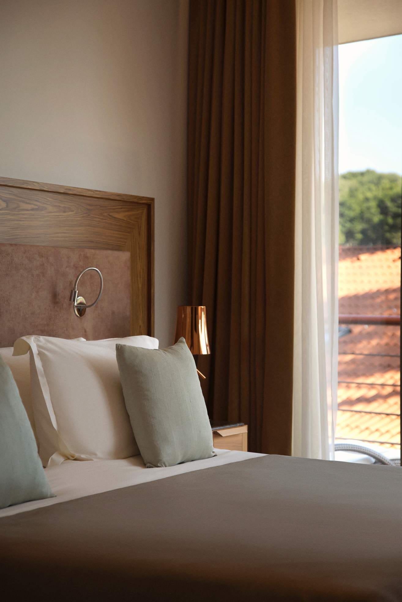 Comfortable bed with soft pillows beside a window with draped curtains, with terracotta rooftops visible outside