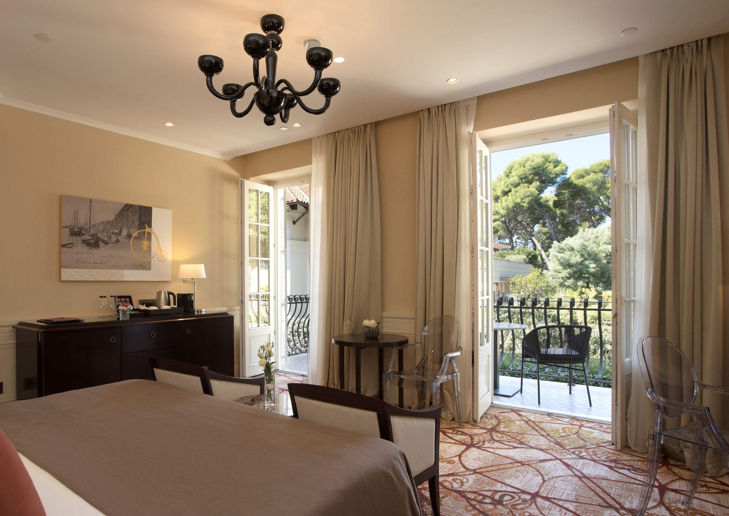 A deluxe room featuring a bed, a desk area, an elegant light fixture, and French doors opening to a balcony with park side views