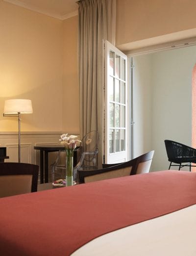 Spacious room with a large bed topped with a red throw, a desk with a lamp, and an open window allowing natural light