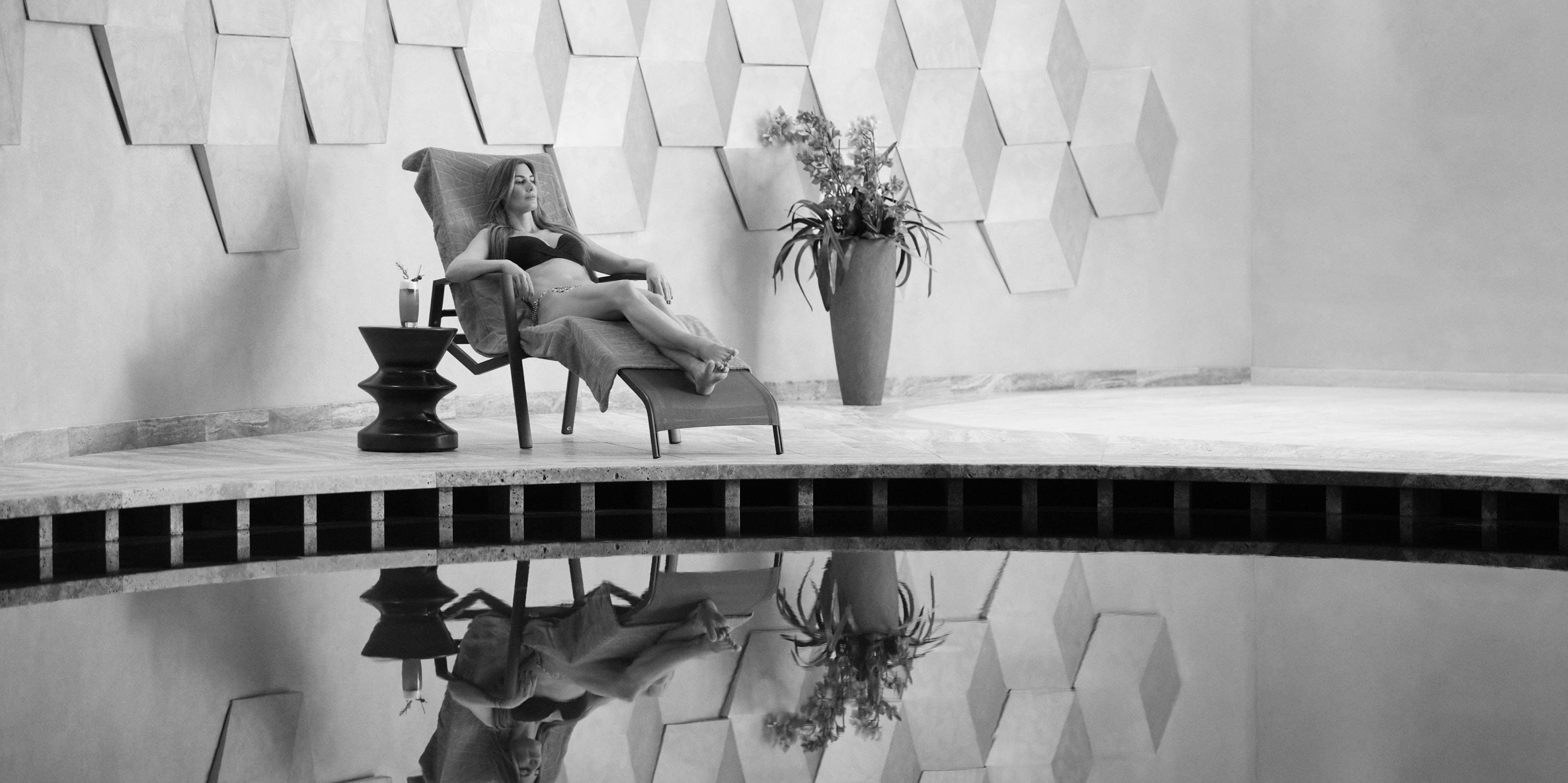 A black and white image of a relaxed woman lounging on a chair by a reflective pool, with a drink on a side table and geometric wall art in the background