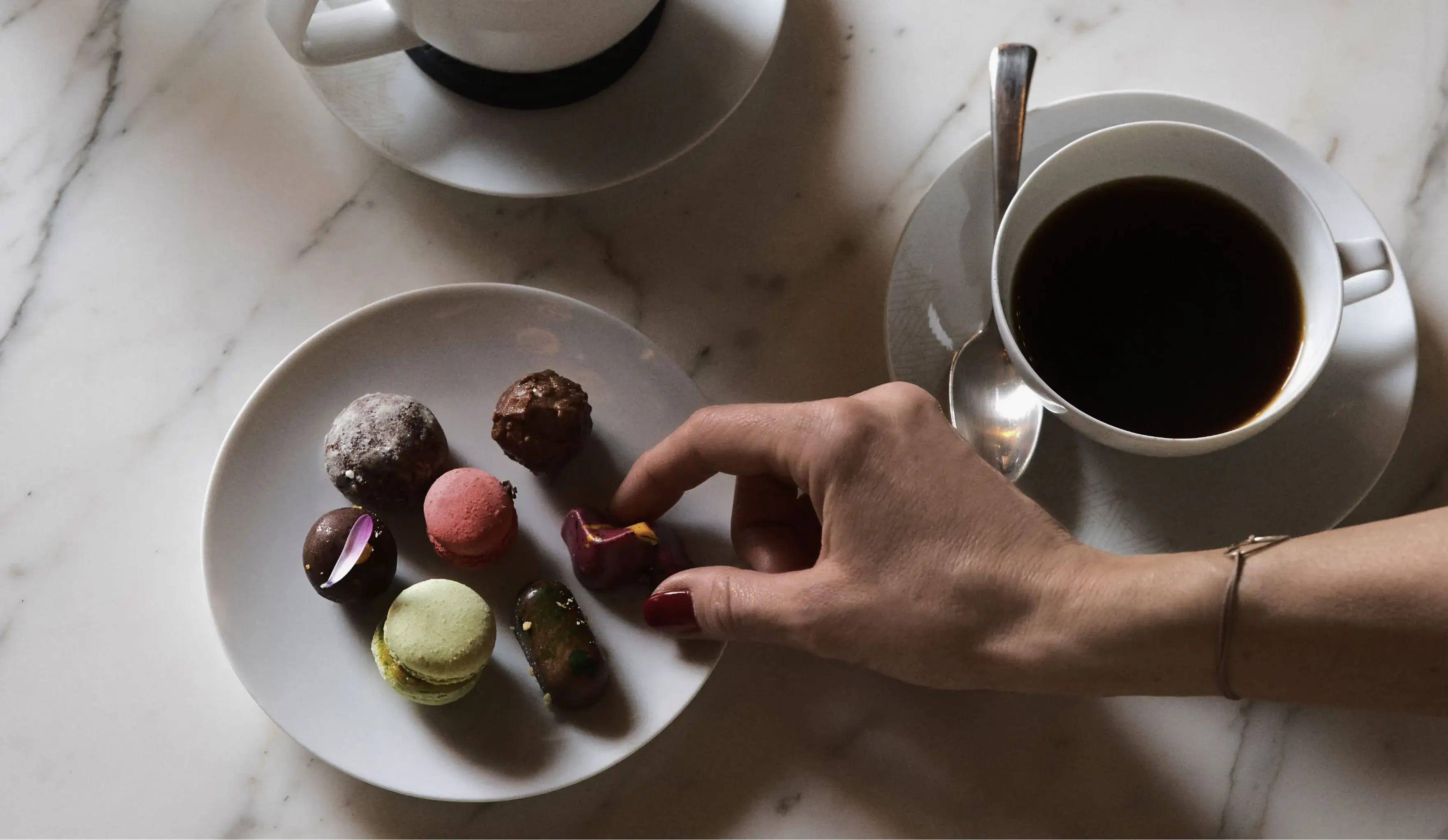A hand selecting a treat from a plate of macarons next to a cup of coffee and a creamer on a marble table