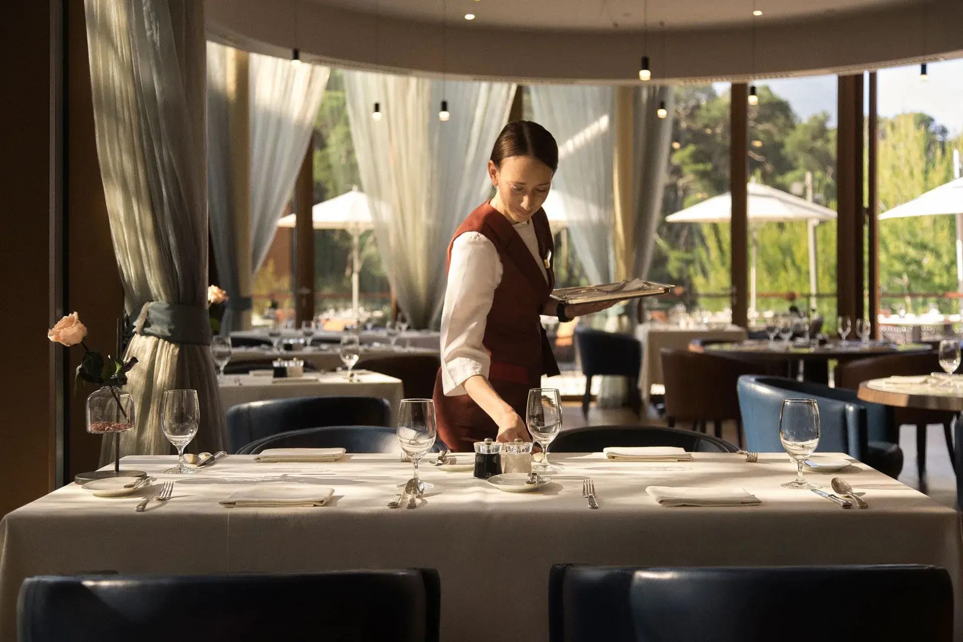 A waitress setting a table in an elegant dining room with large windows and soft natural light