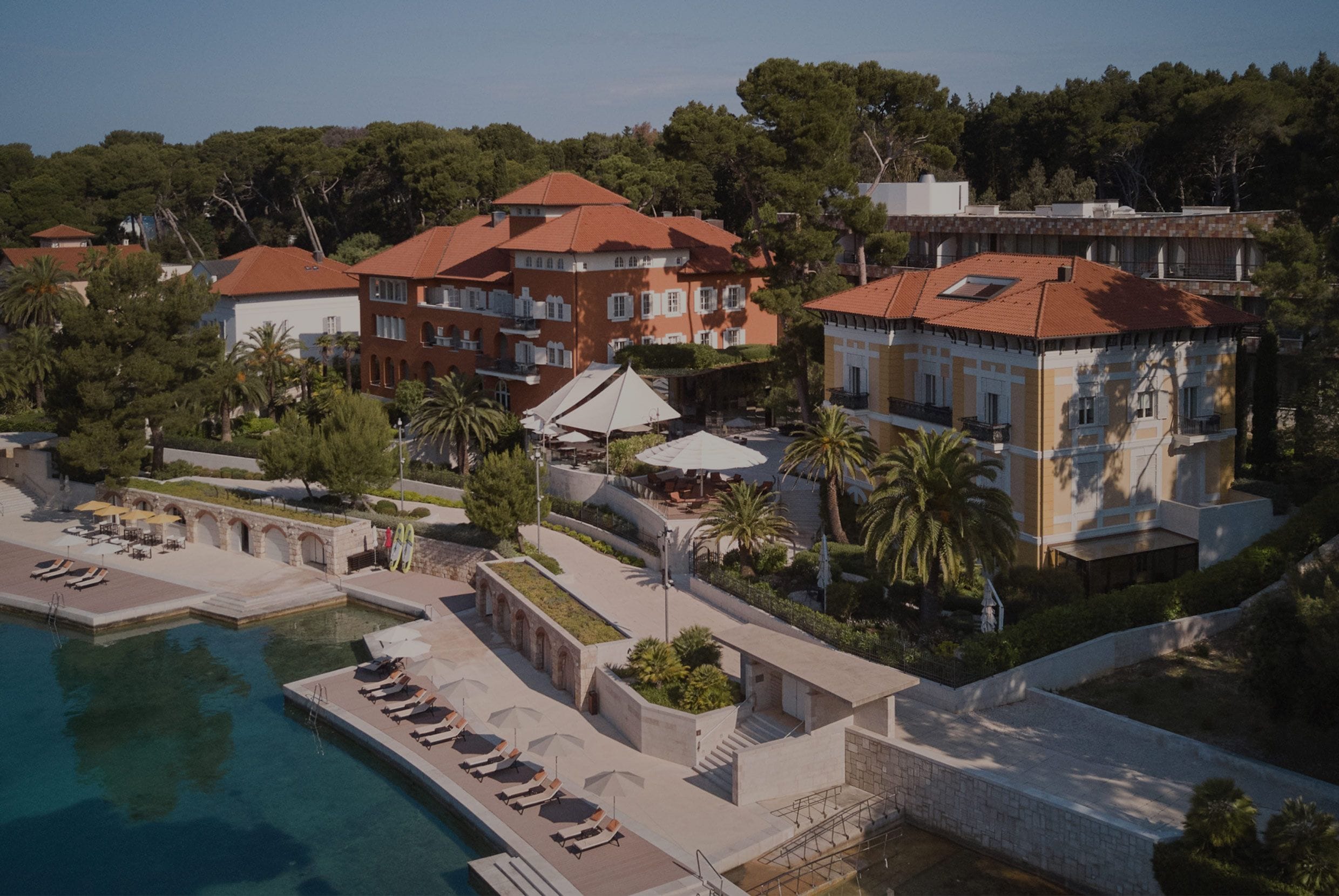 Aerial view of a luxurious resort complex, showcasing terracotta-roofed buildings amid lush greenery, an inviting pool area, and a hint of the serene blue sea in the distance