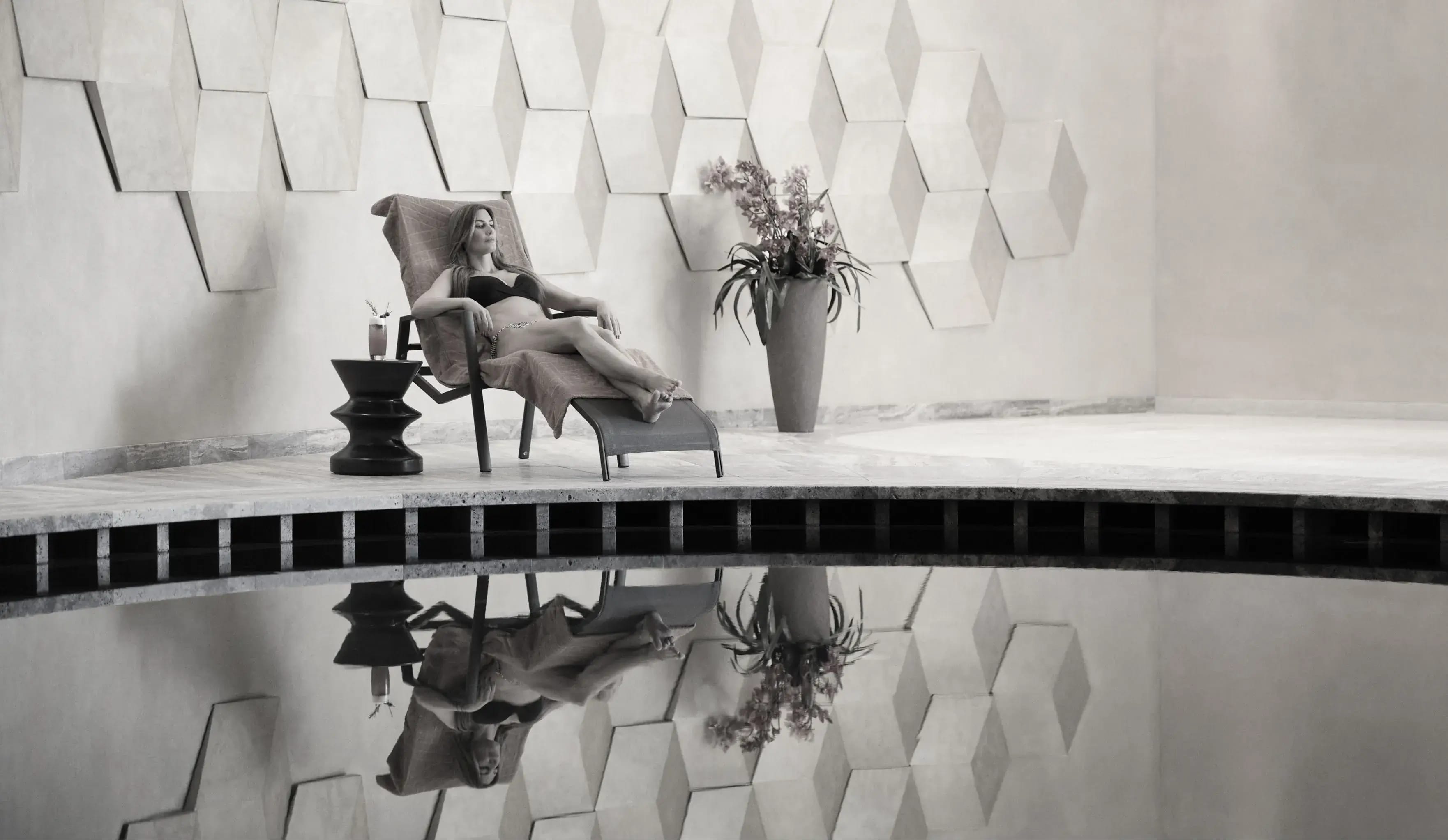 A black and white image of a relaxed woman lounging on a chair by a reflective pool, with a drink on a side table and geometric wall art in the background
