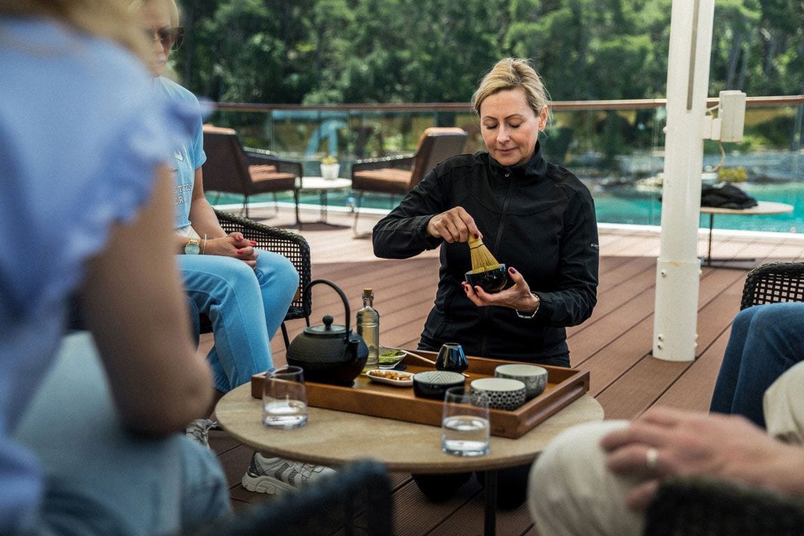 Person preparing tea with a whisk during an outdoor tea ceremony, with several people seated nearby