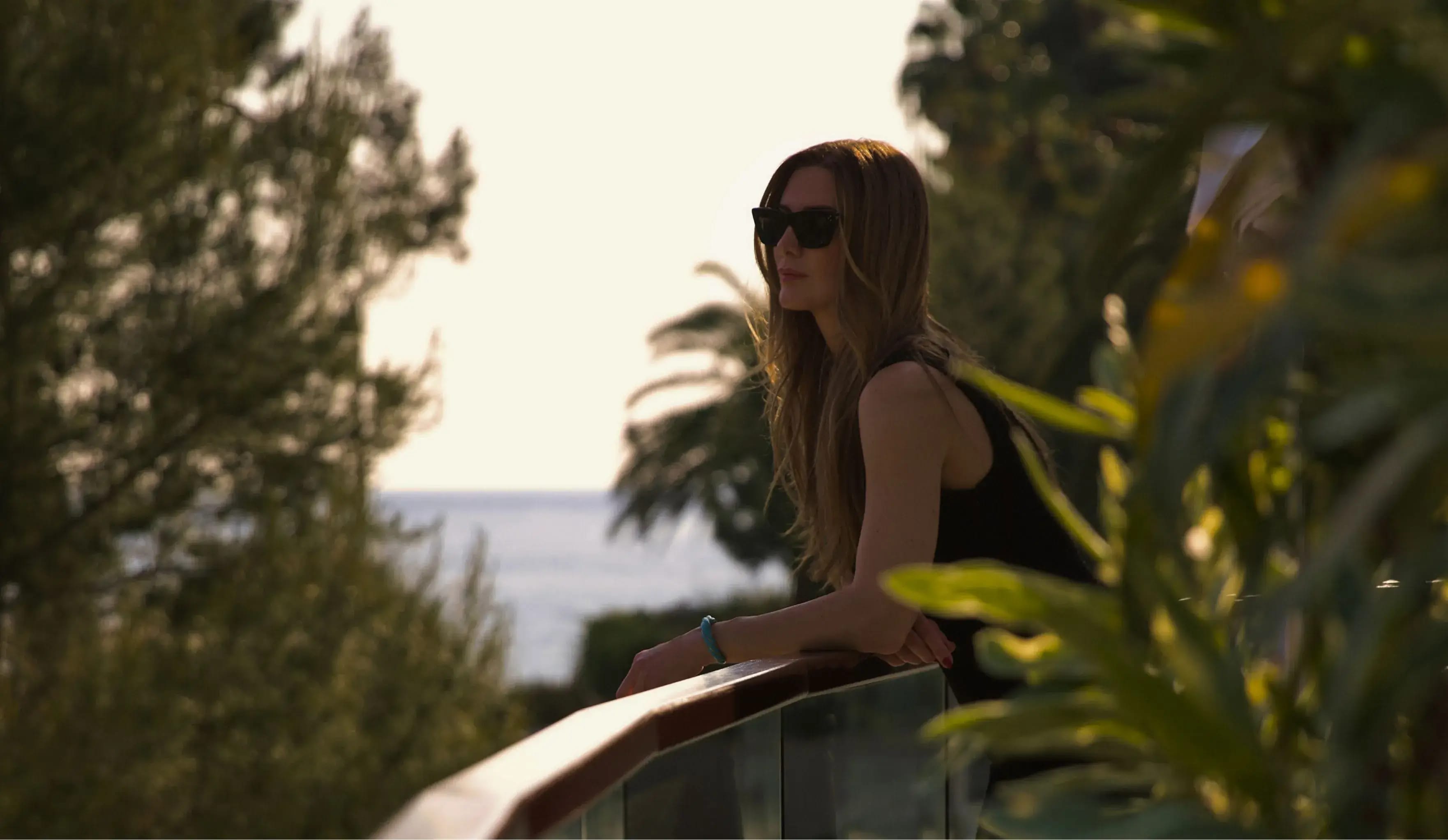 A woman in a casual outfit leaning on a glass railing, surrounded by lush greenery, with a glimpse of the sea in the background
