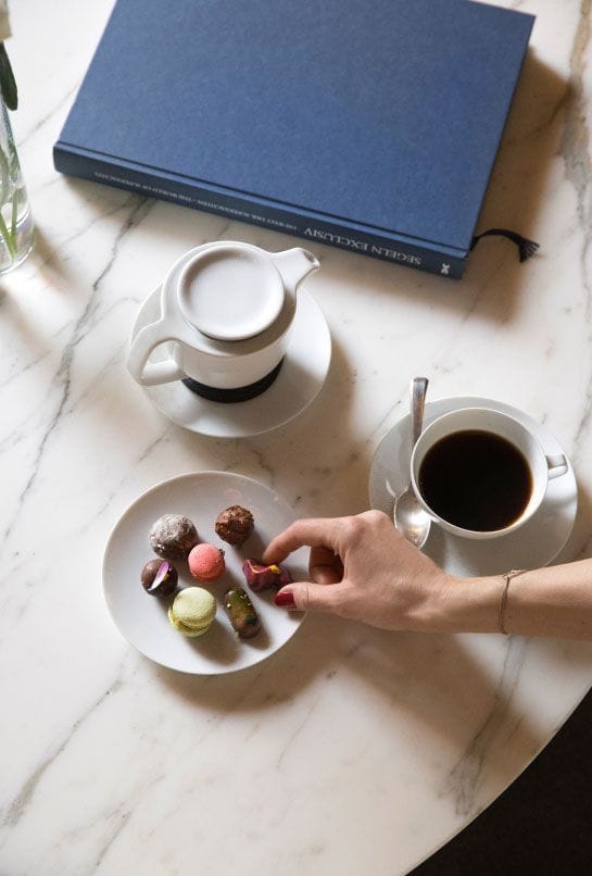 A hand selecting a treat from a plate of macarons next to a cup of coffee and a creamer on a marble table