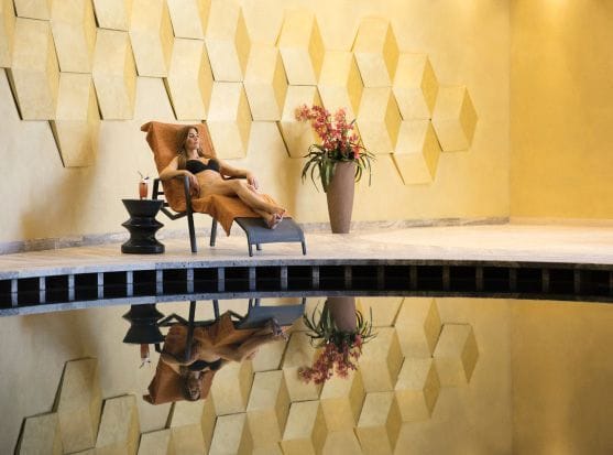 A relaxed woman lounging on a chair by a reflective pool, with a drink on a side table and geometric wall art in the background