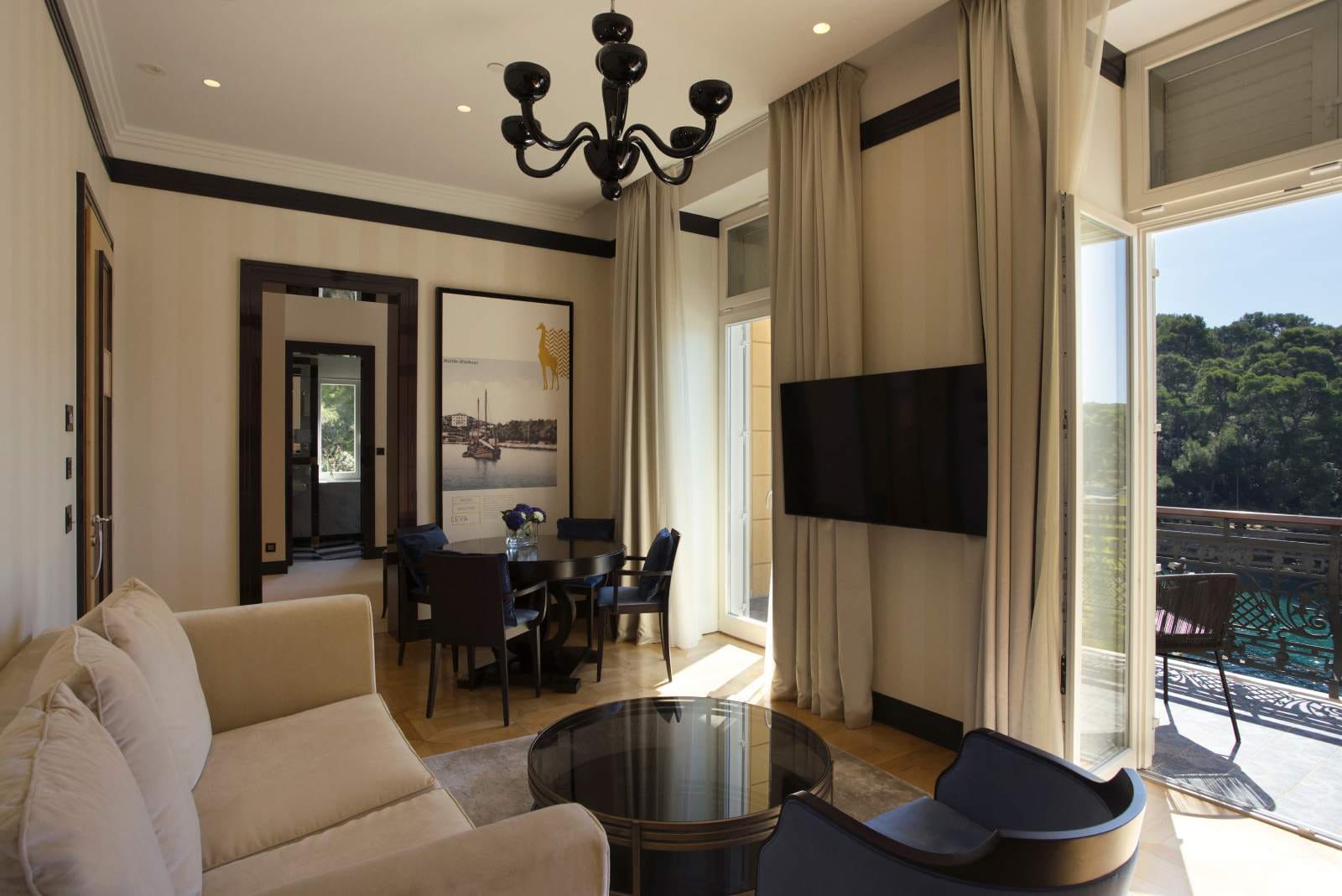 A chic, spacious suite featuring a plush sofa, a glass-top coffee table, and a dining area leading to a balcony with a scenic view
