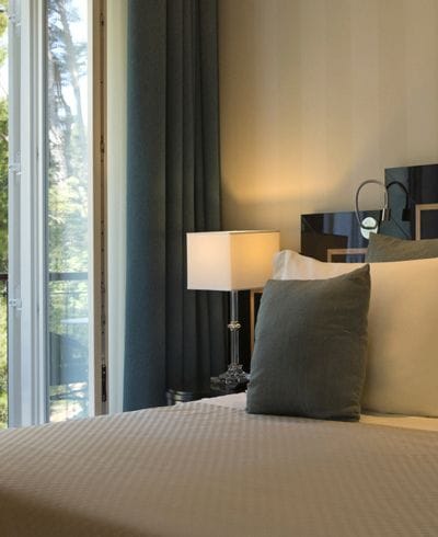 A modern bedroom with a large bed, elegant lamps, and a view of lush greenery through full-length windows