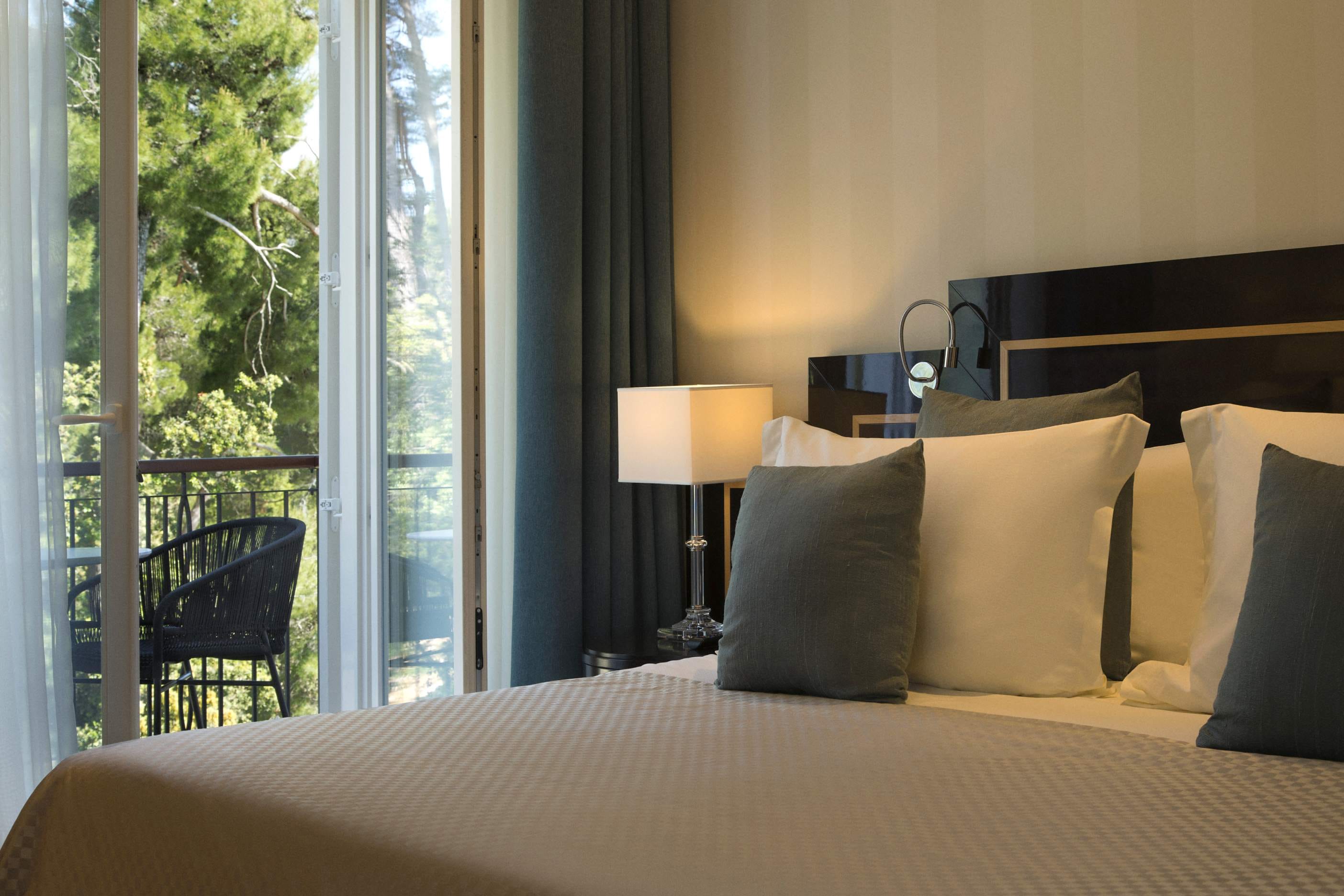 A stylish bedroom featuring a comfortable bed with plush pillows, chic lighting, and a balcony with a serene view of tall trees