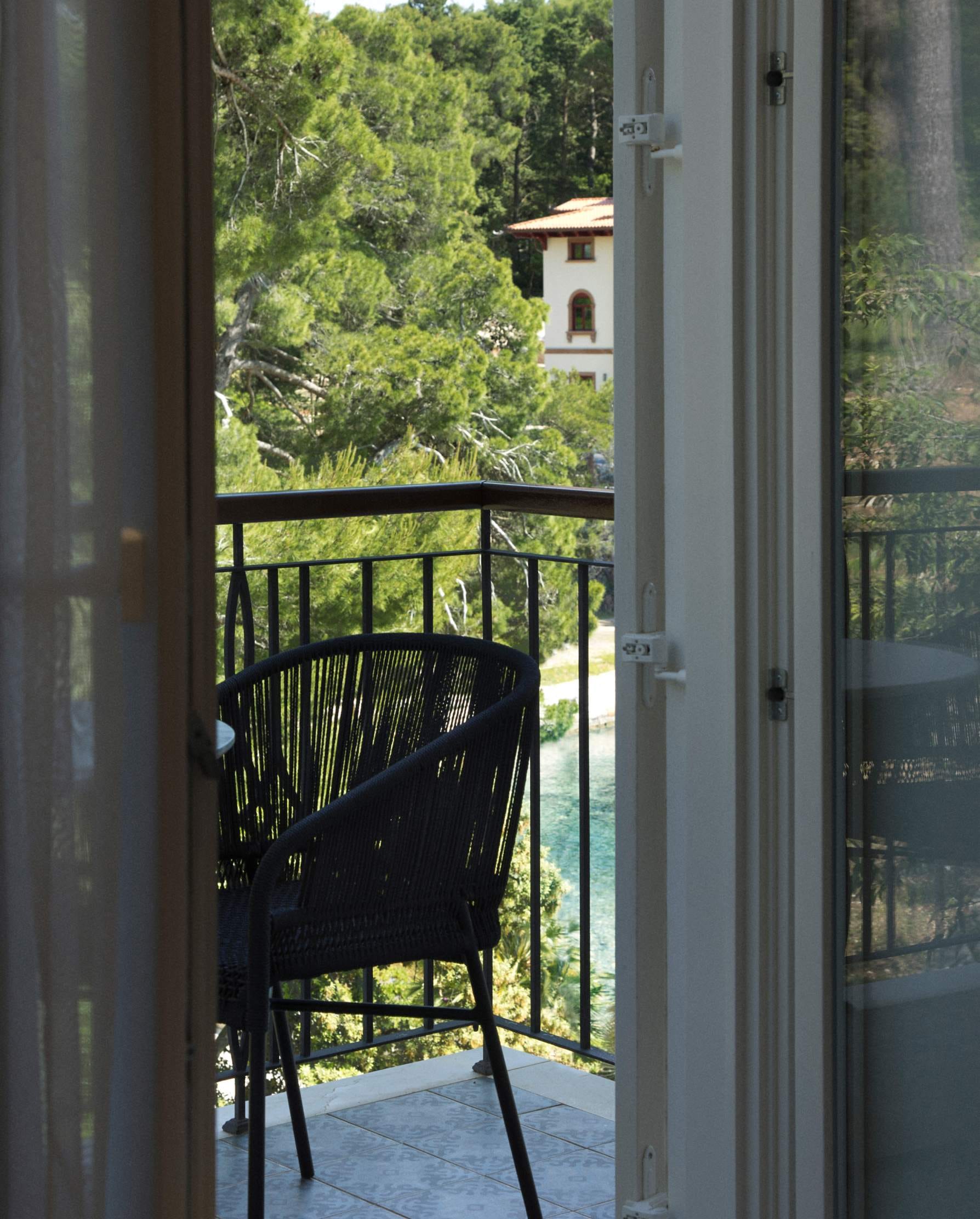 An open door leads to a balcony with a wicker chair, overlooking lush greenery and a Mediterranean-style building