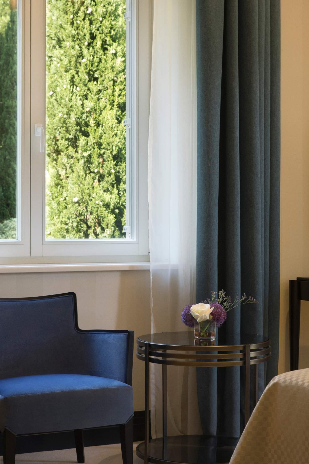 A cozy corner with a blue armchair and a small round table holding a flower vase, beside a window with a view of green foliage