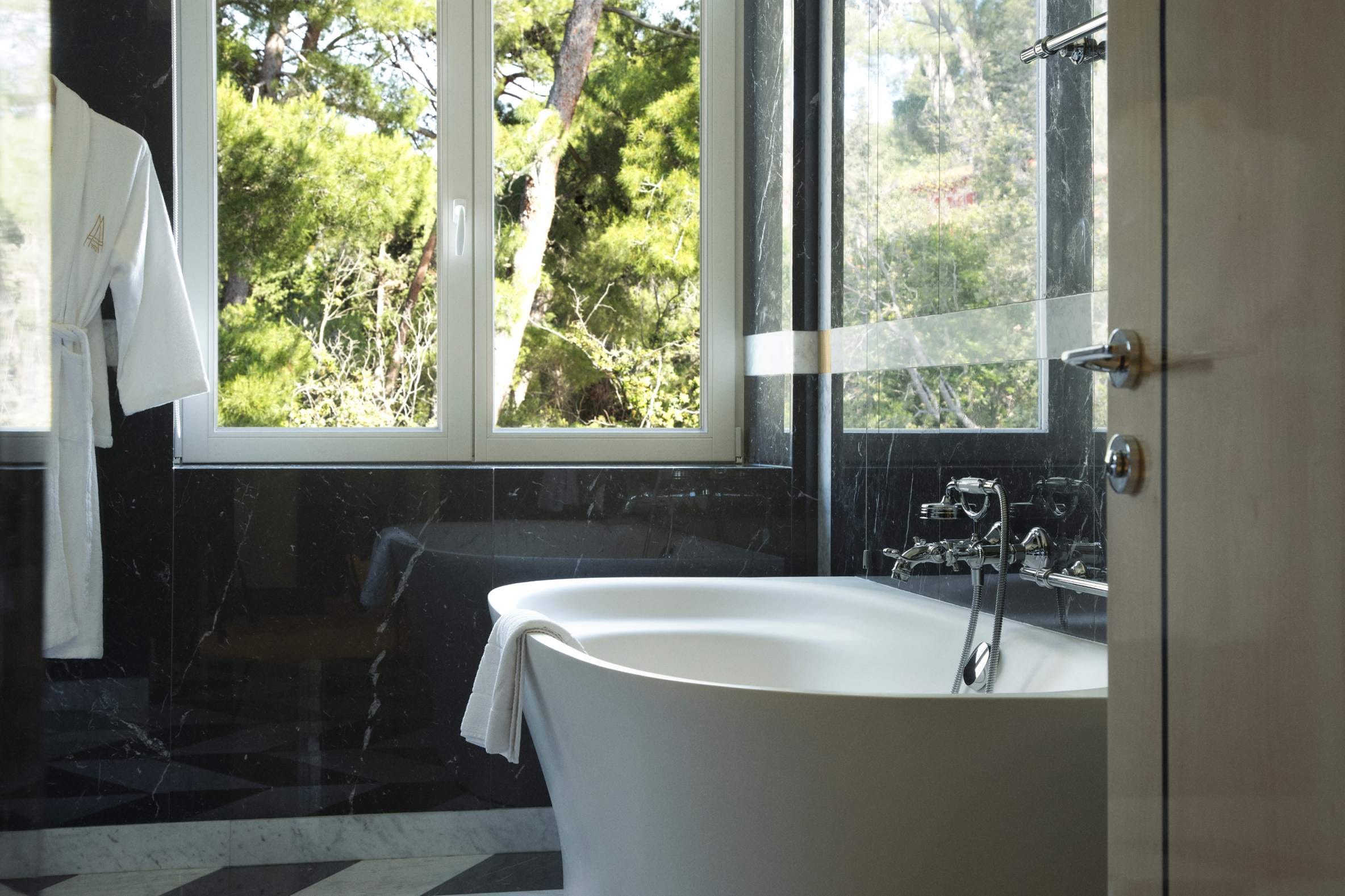 An elegant bathroom with a standalone bathtub, black marble walls, and a window offering a view of lush trees