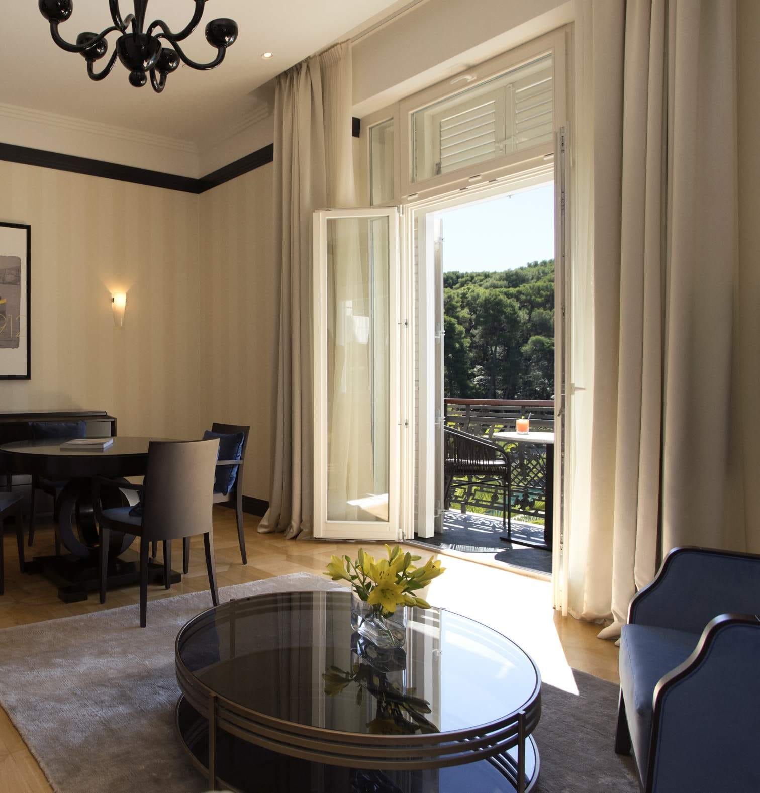 An elegant room with an open balcony door, a round glass table in the foreground, and a dining area in the background bathed in natural light.