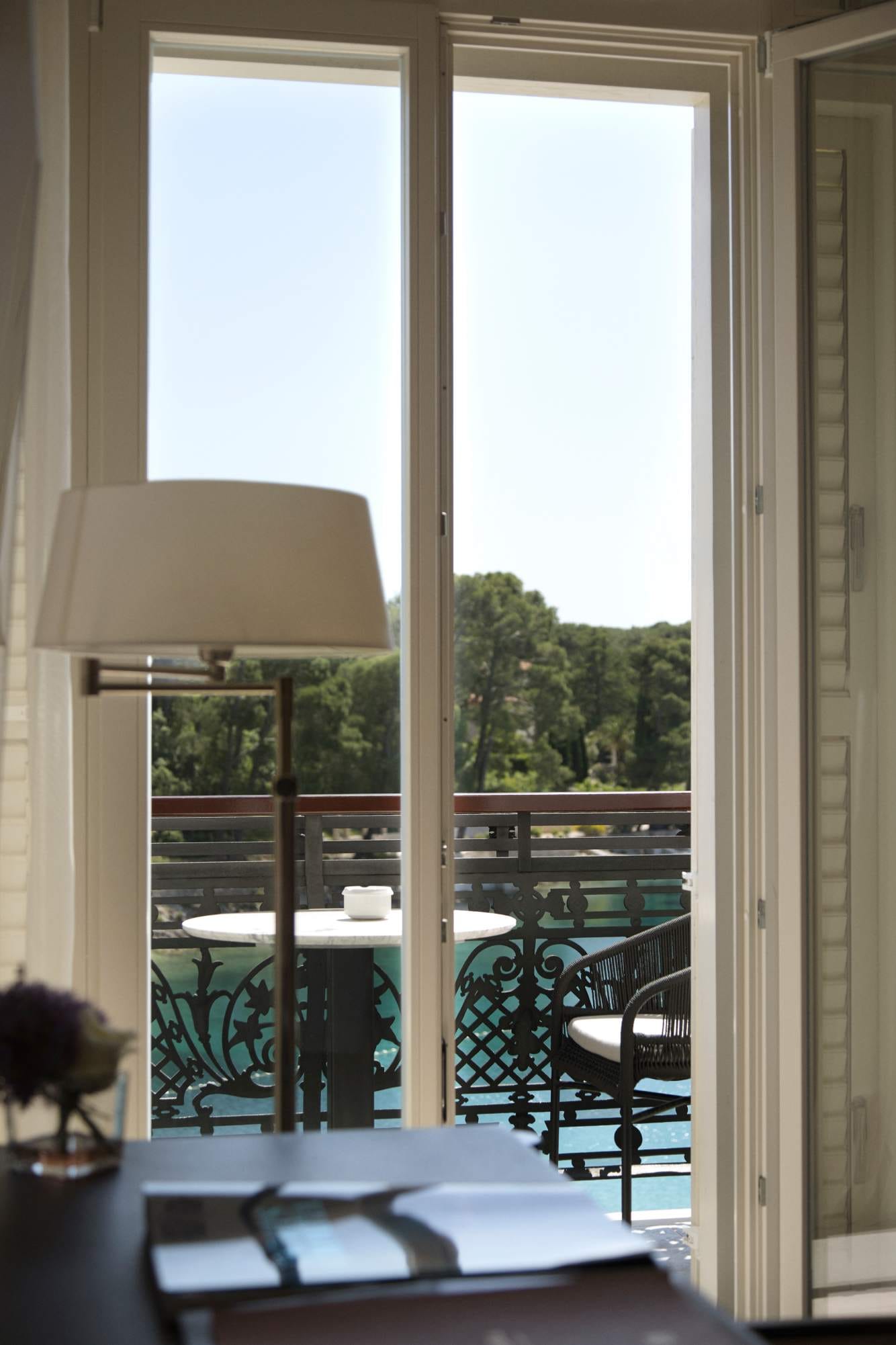 A view through an open door leading to a balcony with outdoor furniture, framed by sheer curtains and a room lamp in the foreground.