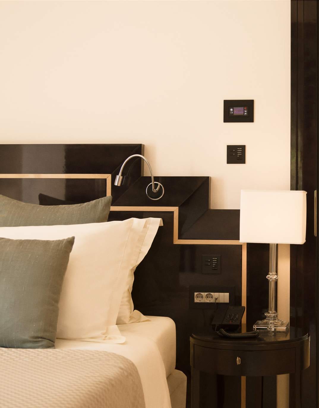 A bedside area with a modern lamp and reading light, plush pillows against a dark headboard, and an elegant side table with a phone and room controls