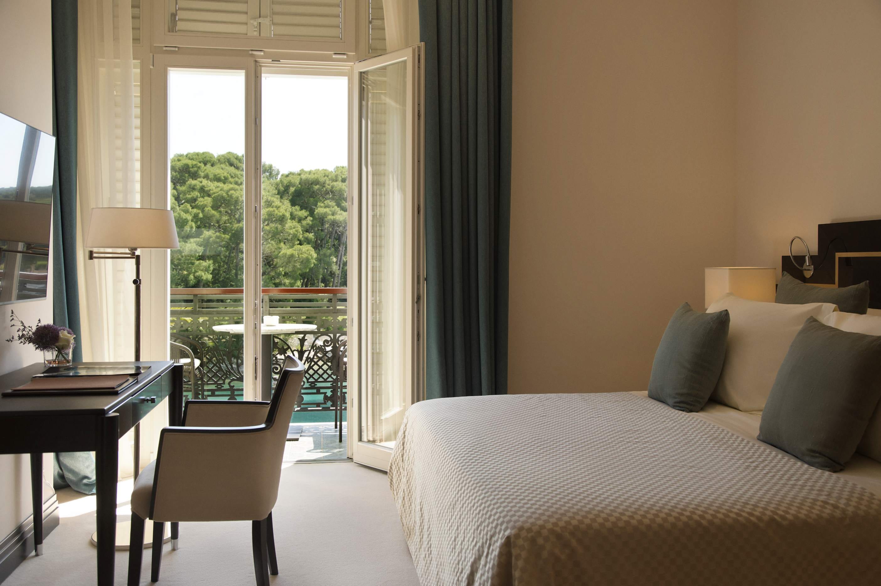 A warm and inviting bedroom with an open balcony, a desk with a lamp, and a comfortable bed adorned with soft pillows, bathed in natural light