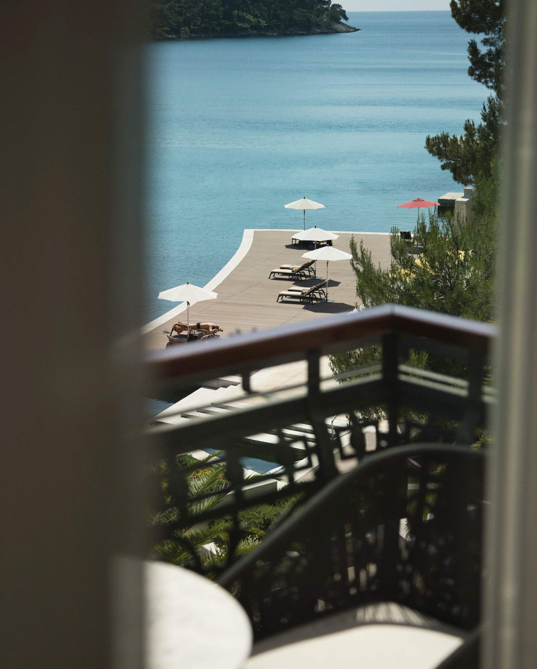 A peek through balcony railings at a serene seaside lounging area with umbrellas and sunbeds, overlooking calm blue waters
