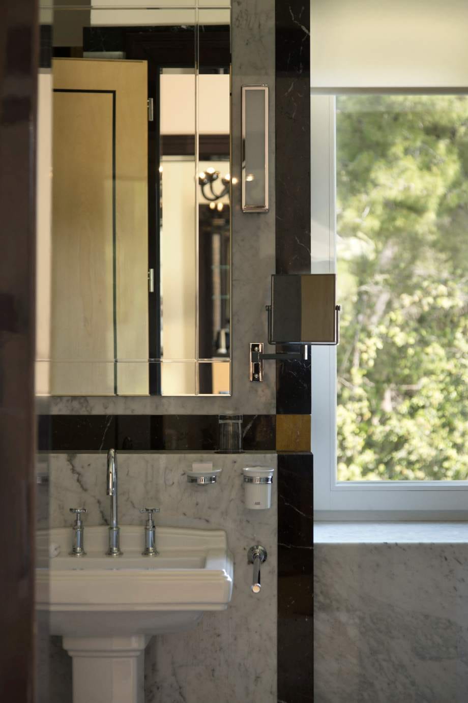 A luxurious bathroom reflected in a mirror, showcasing a sleek sink set against marble walls, with a view of greenery through the window.