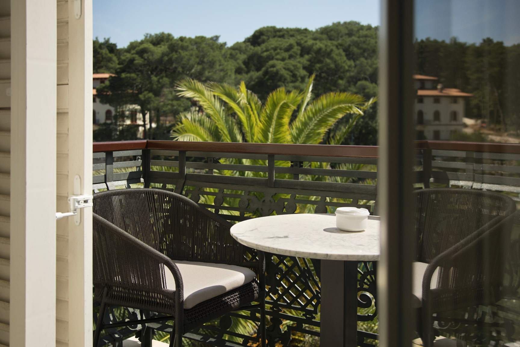 A cozy balcony setup with a marble table and wicker chairs, overlooking lush palm trees and a serene landscape