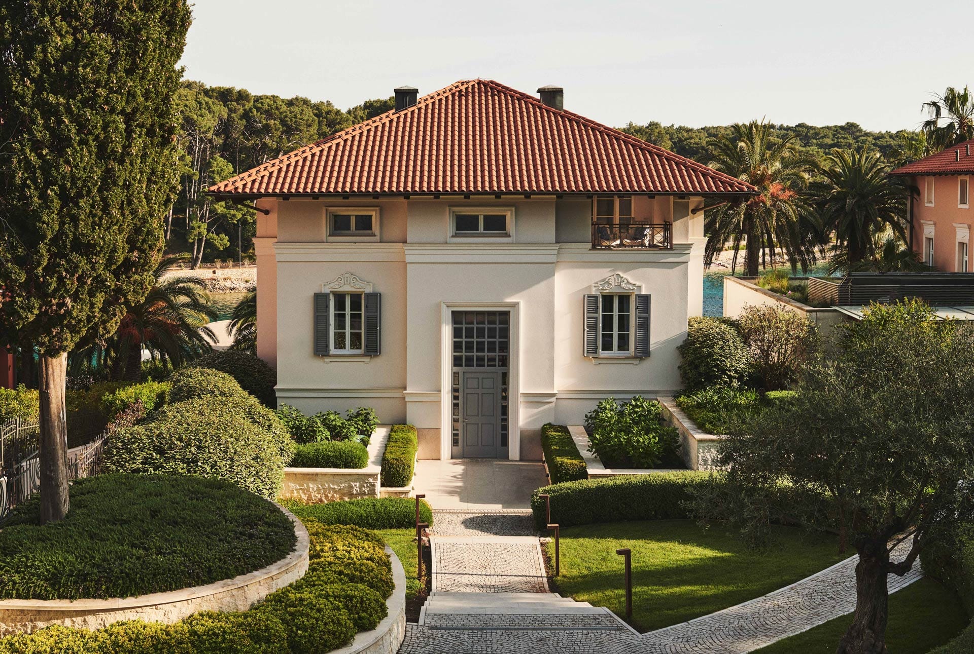 A well-manicured villa with a tiled roof, flanked by lush greenery and overlooking a beach with palm trees.