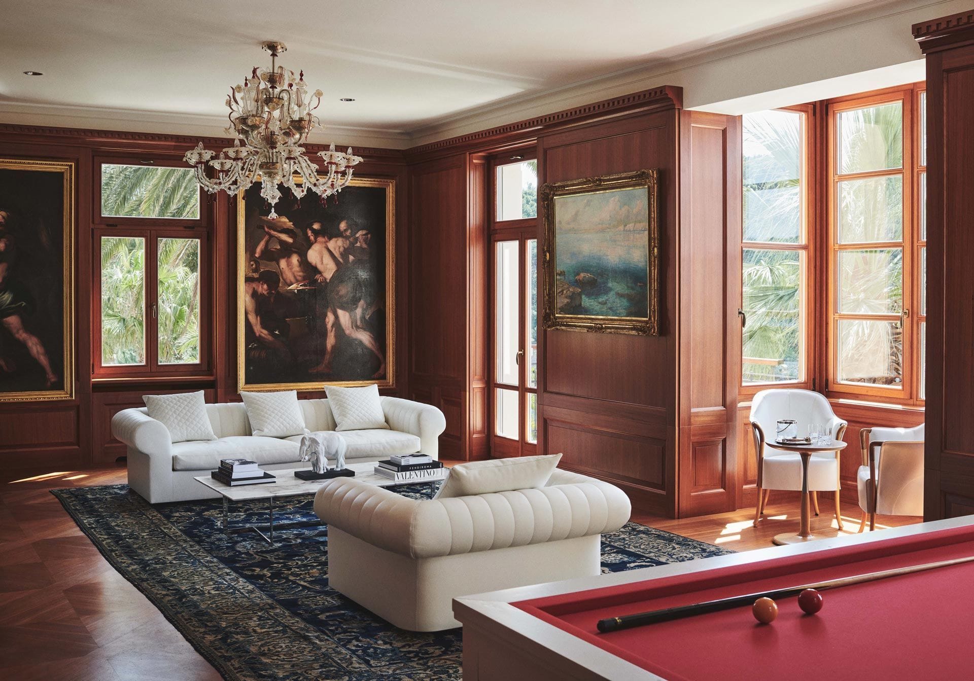 Classic living room with a white sofa, billiards table, ornate chandelier, and framed artwork, with sunlight streaming in through window shutters.
