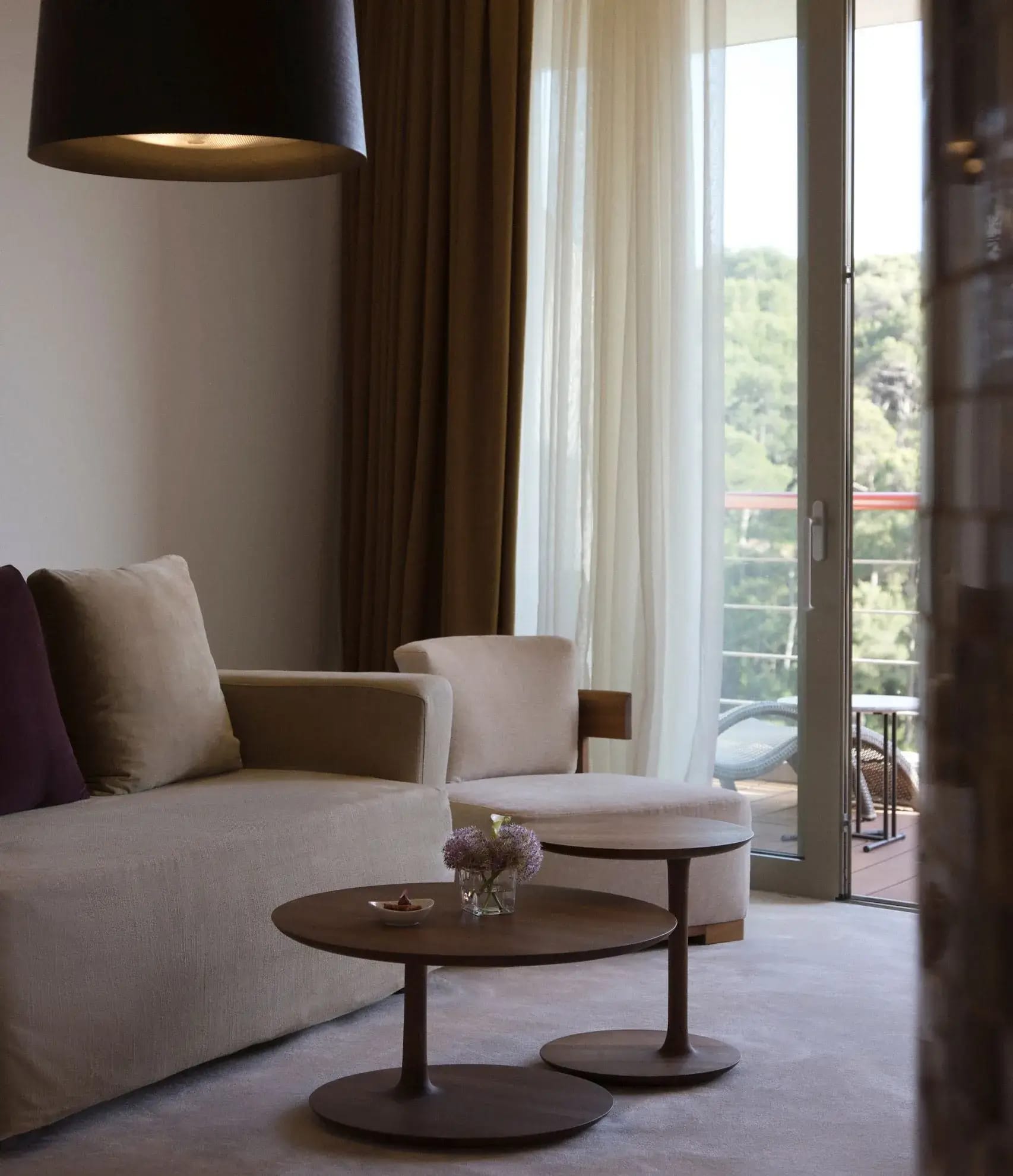 Interior of a hotel room with a beige sofa and armchairs, wooden coffee tables, and sheer curtains, leading to a balcony with a green view.